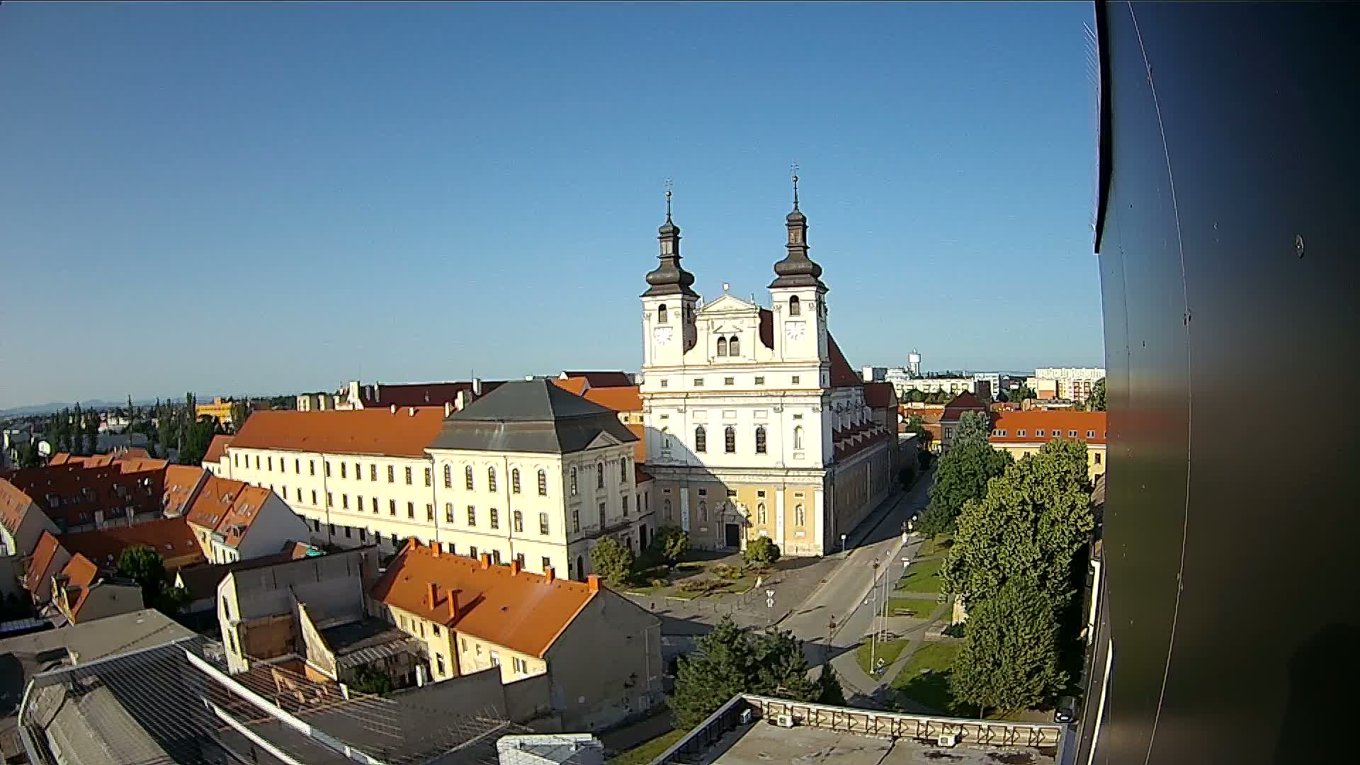 An elevated view shows a large, light-colored church with two towers in the center of a town on a clear, sunny day.