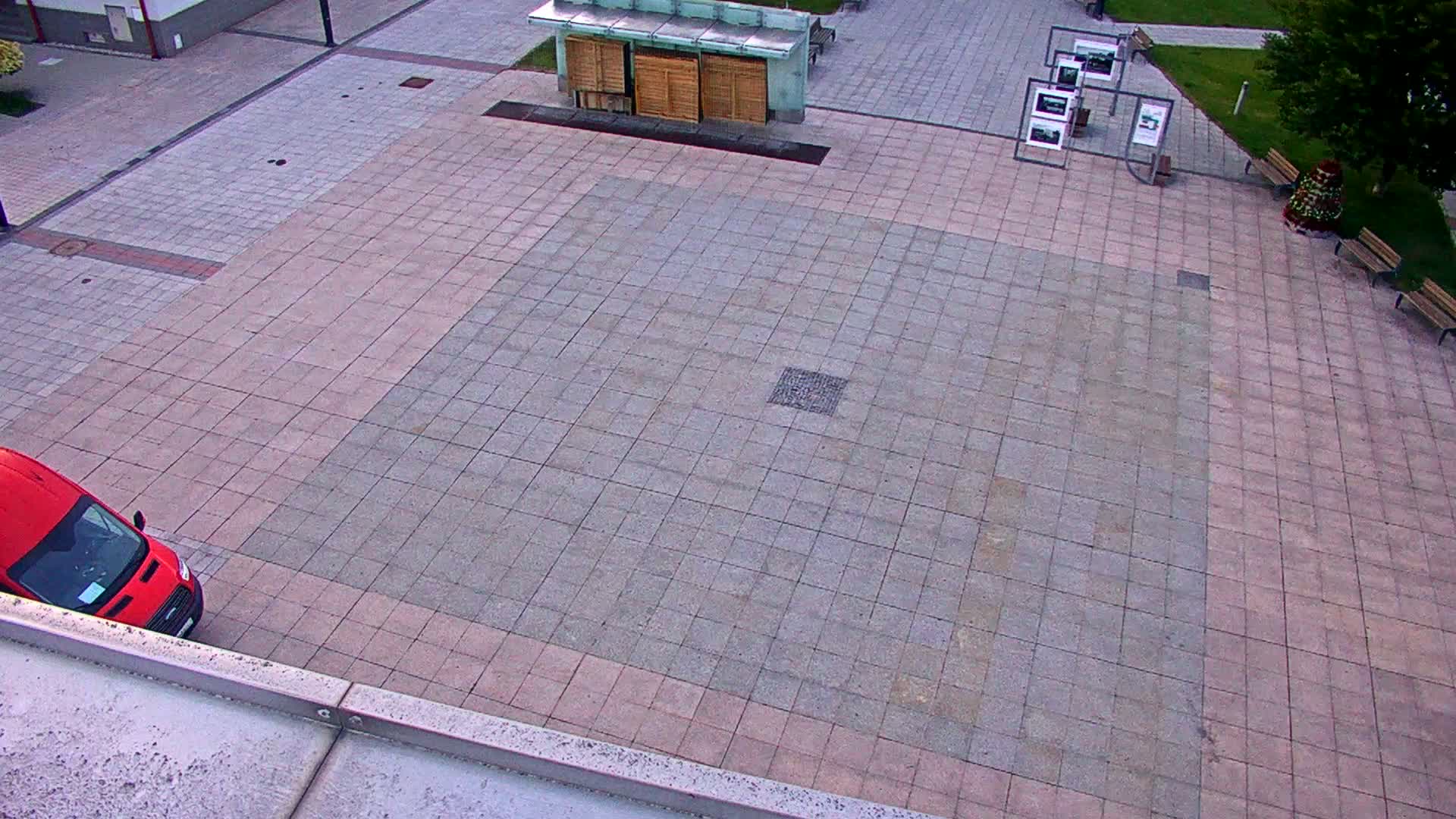 A red van is parked in a large paved plaza with a small kiosk and several benches, under an overcast sky.