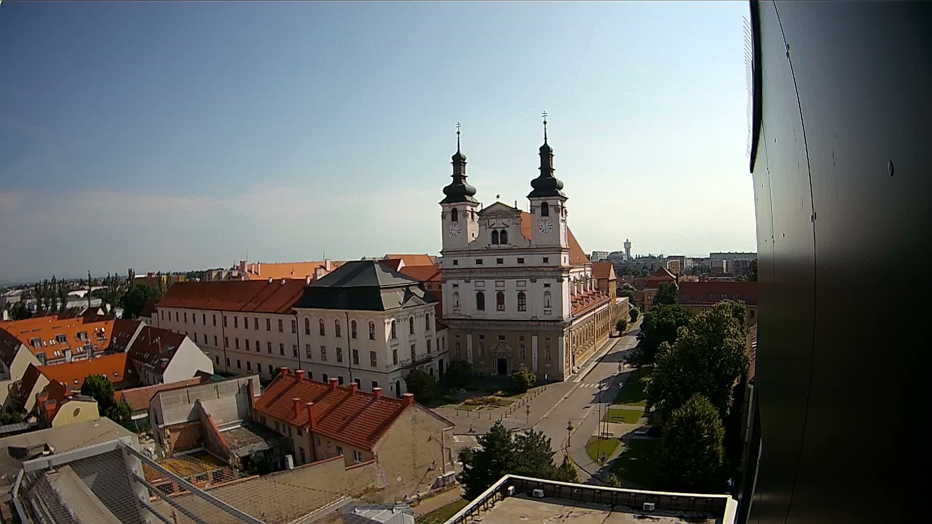 A high-angle view on a clear, sunny day shows a large church with two towers at the center of a town square surrounded by low buildings with red tile roofs.