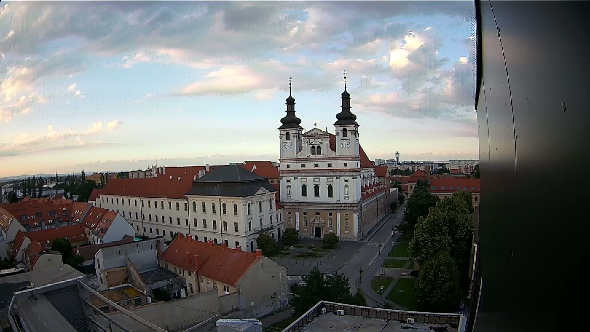 An elevated view of a city under a partly cloudy sky shows a large church with two towers at the center, surrounded by other buildings.