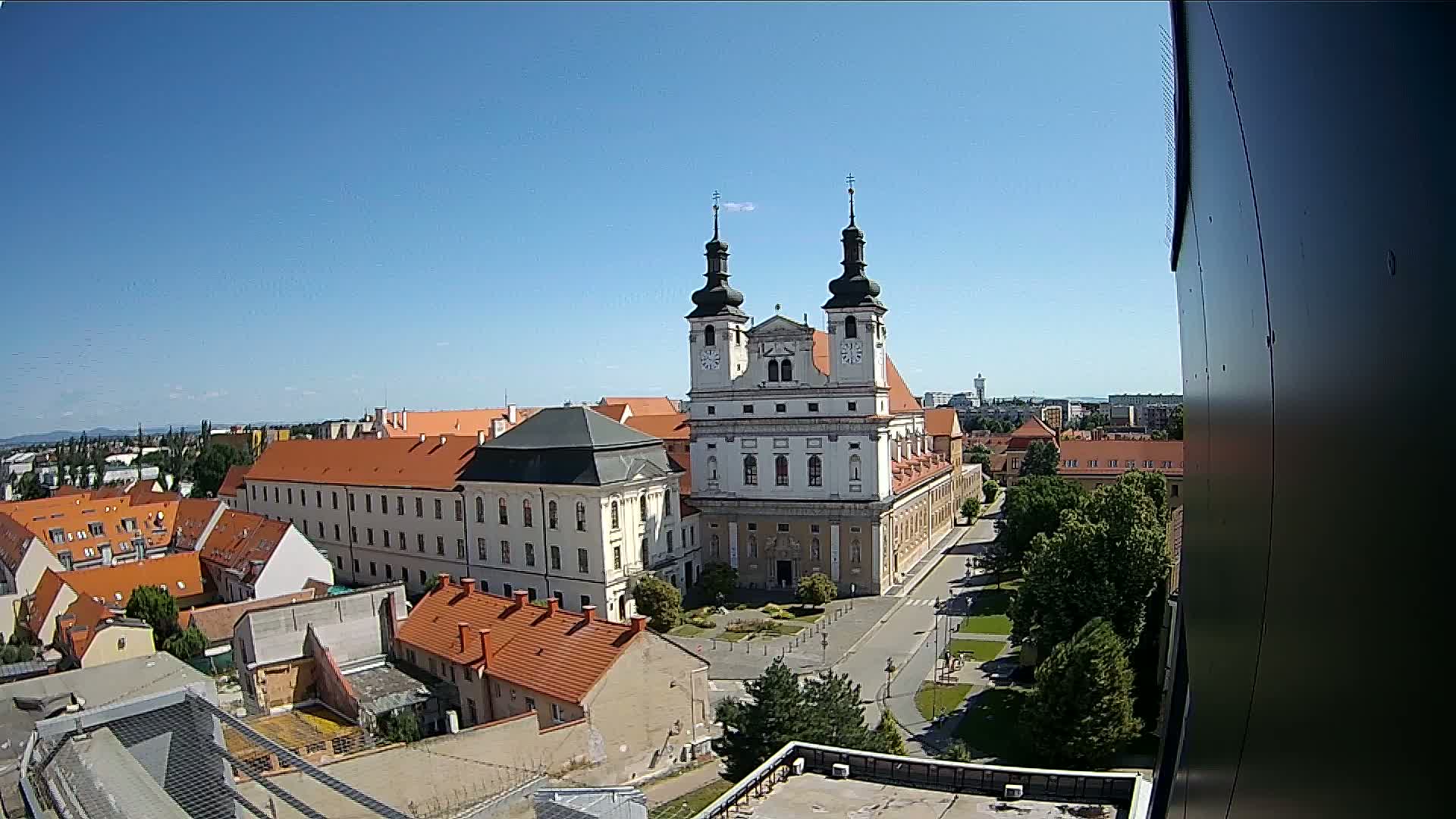 A large white church with two towers stands prominently in a town square, surrounded by low-rise buildings with red-tiled roofs under a clear blue sky.