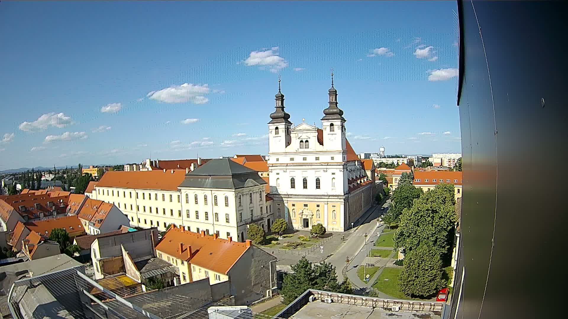 A large, pale yellow church with two dark towers stands in a city square on a sunny day with scattered clouds.
