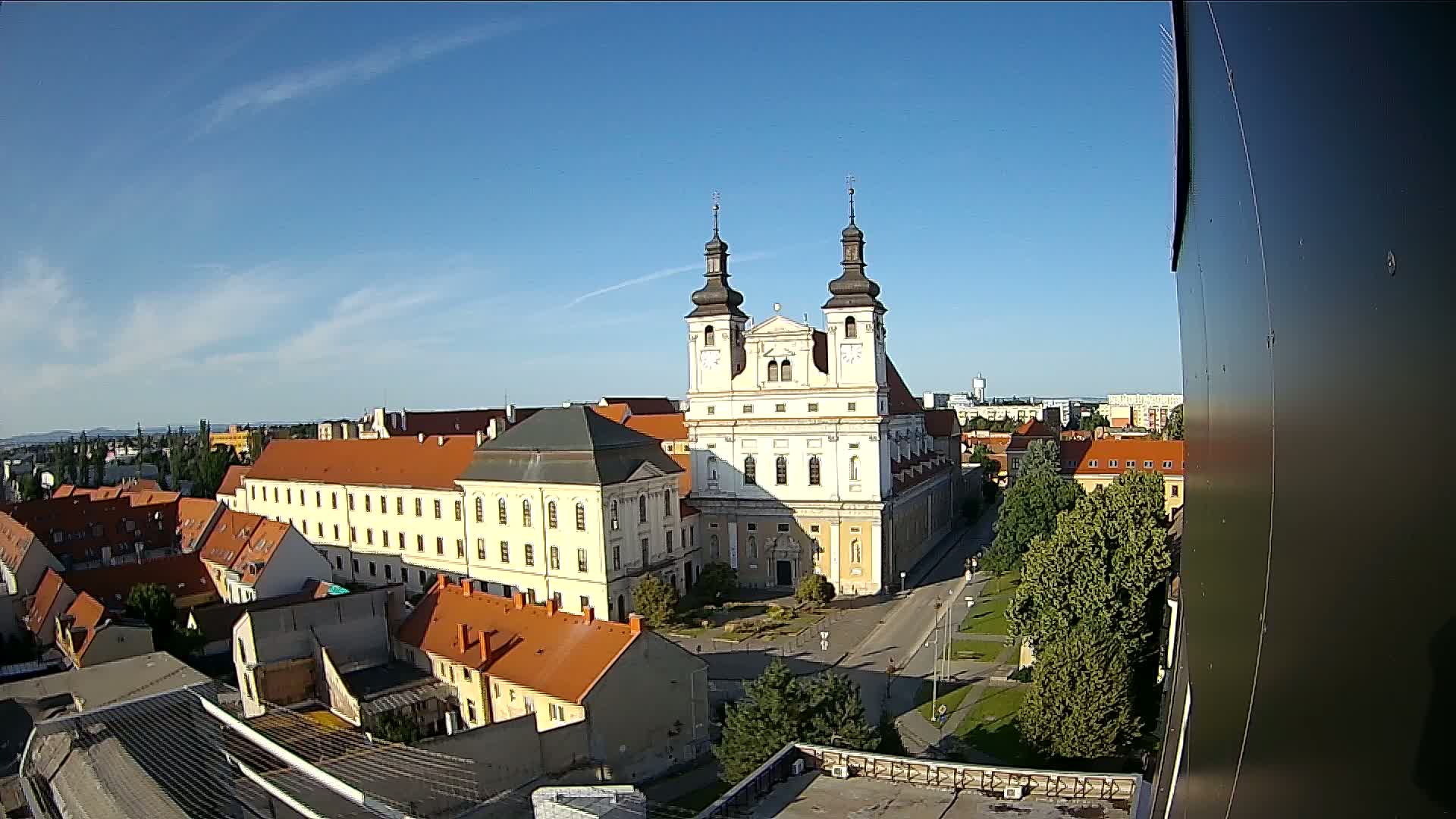 An elevated view on a sunny day shows a large, light-colored church with two towers, surrounded by other buildings with red-tiled roofs, and green trees.