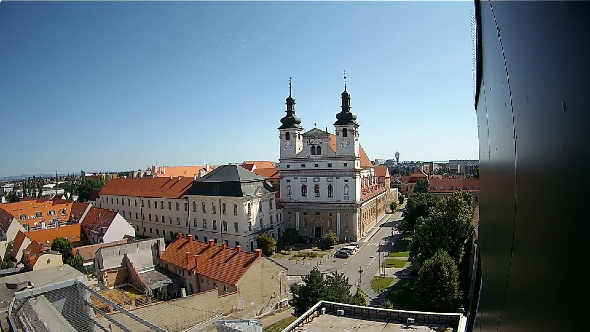 A large church with twin towers stands in a city square on a sunny day, surrounded by other buildings and green spaces.