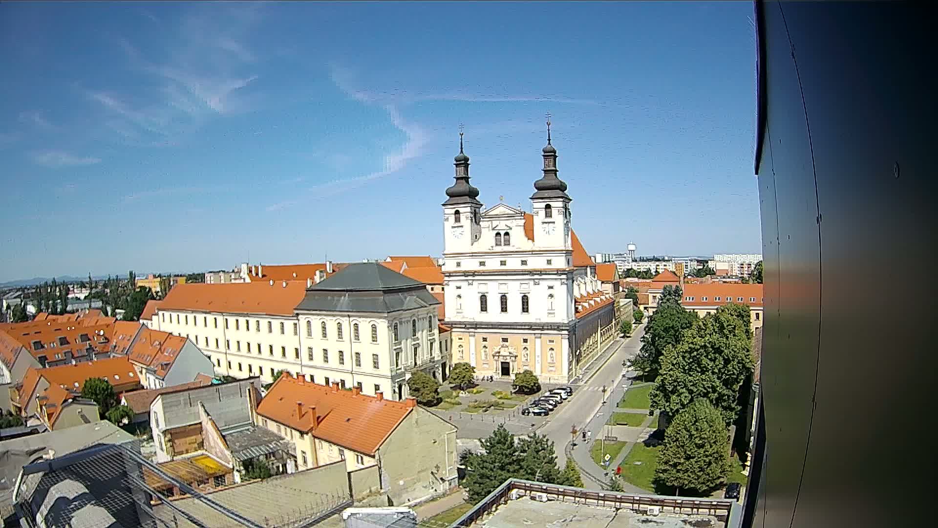 A large church with twin towers stands prominently in a city center under a mostly clear, sunny sky, surrounded by smaller buildings and green spaces.