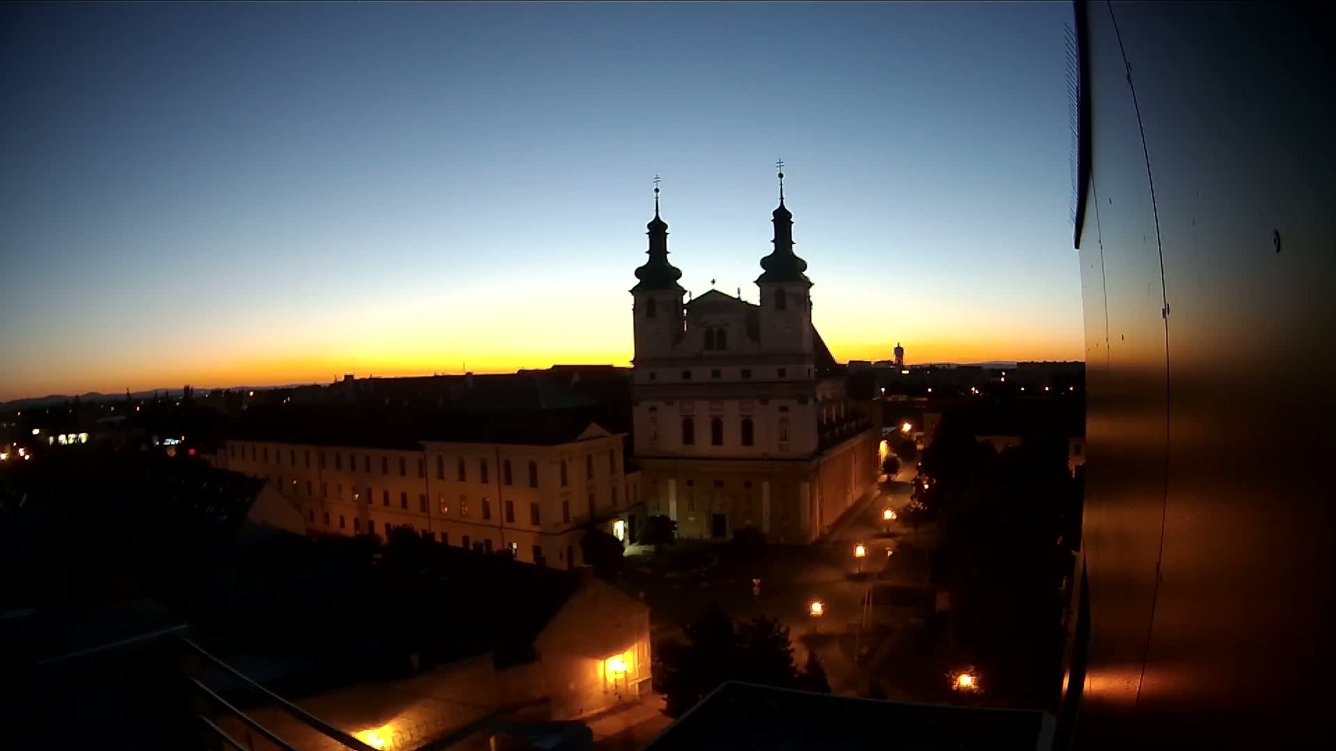 A city at sunset, showing a large church and surrounding buildings illuminated by streetlights.