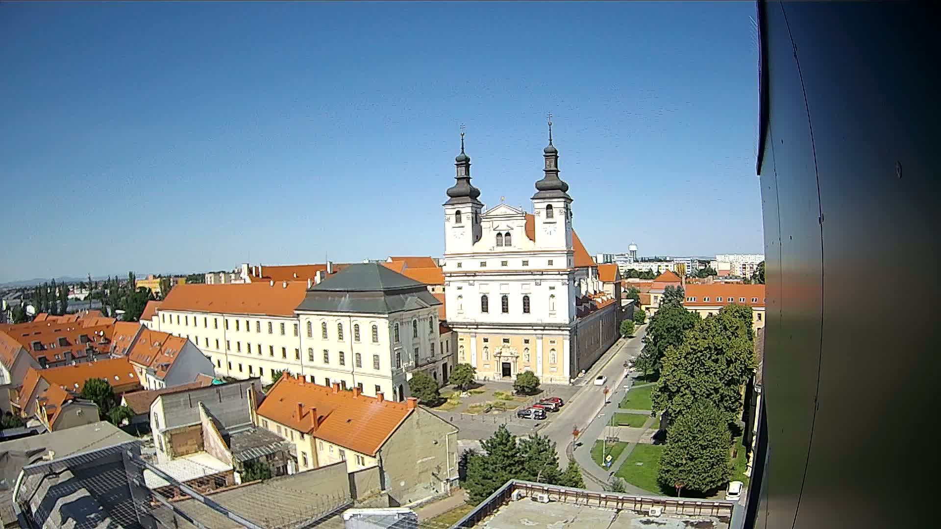 An aerial view on a clear, sunny day shows a large church with twin towers, surrounded by buildings with orange roofs and a small park.