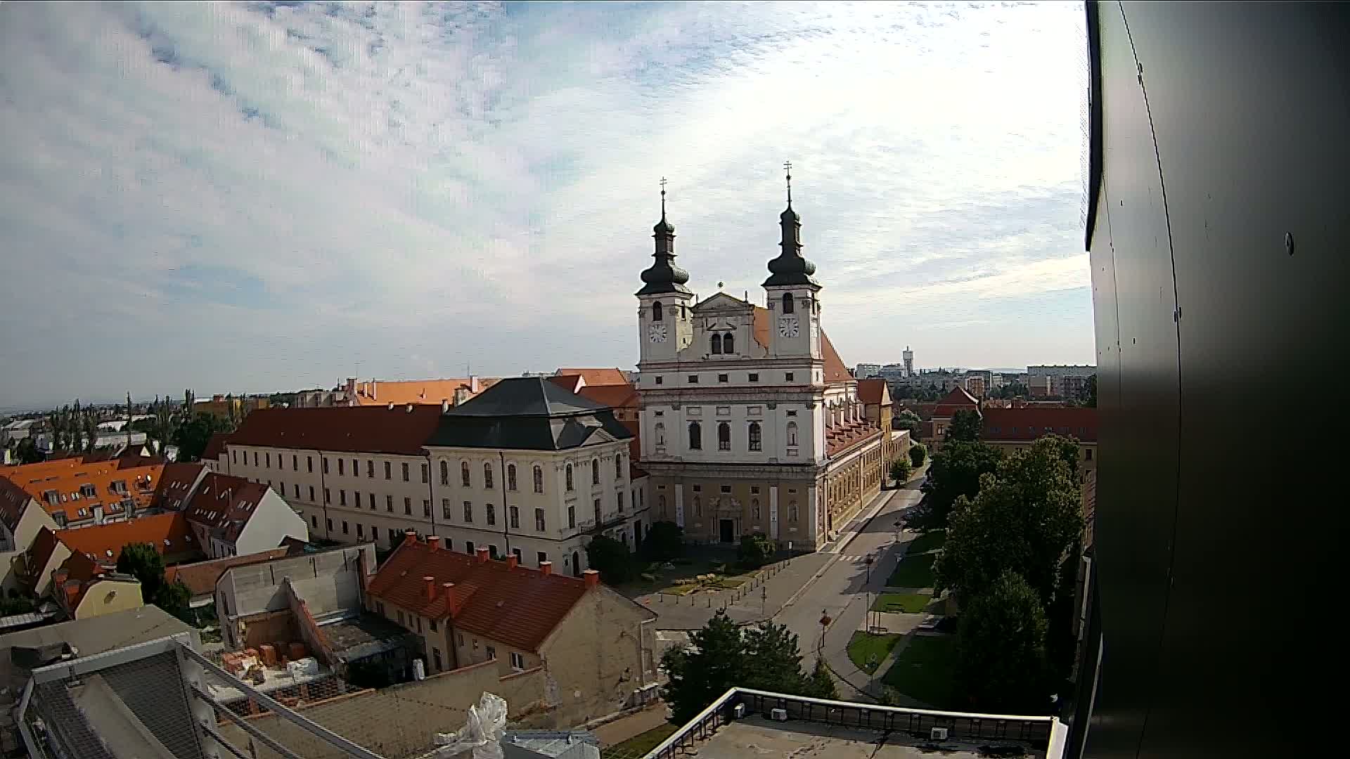 An elevated view of a European city on a partly cloudy day shows a large church complex surrounded by buildings with red-tiled roofs and green trees.