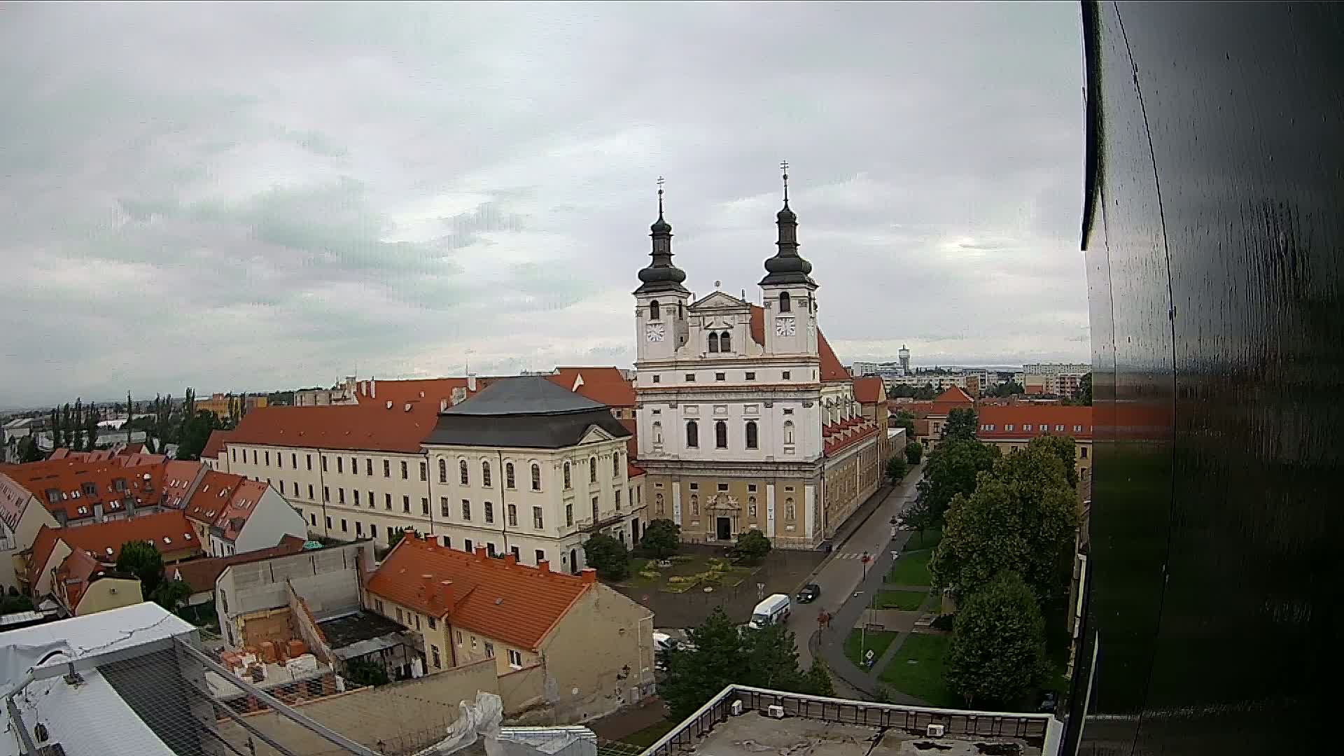 A large church with two towers stands in the center of a city under a cloudy sky.