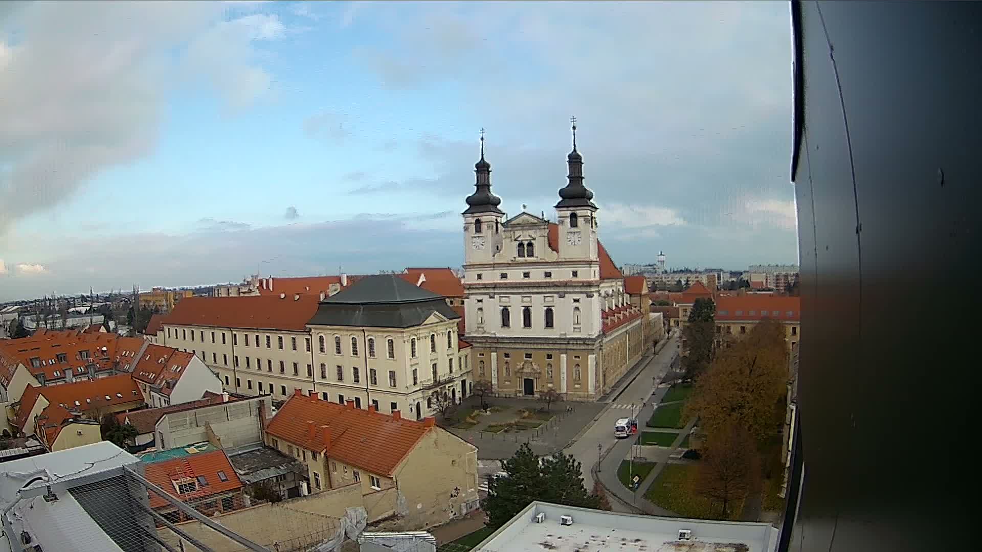 An aerial view of an old European city reveals a grand, two-towered baroque church and an adjacent stately building at its heart, surrounded by numerous red-roofed structures, all under a partly cloudy sky.