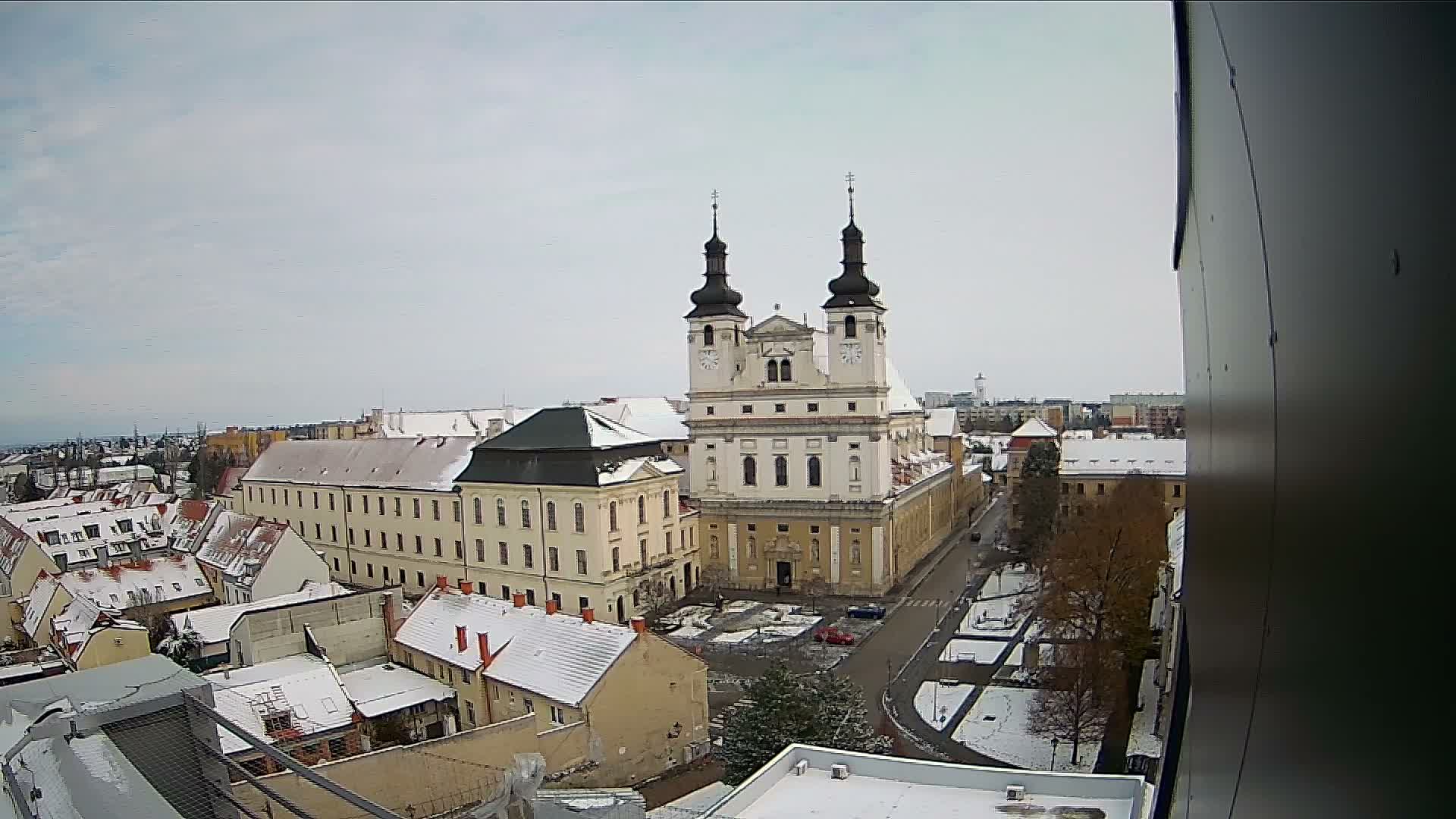 A high-angle view captures a snow-dusted European town, featuring a grand Baroque church with twin spires dominating the scene amidst other buildings and streets, all beneath a bright, overcast winter sky.