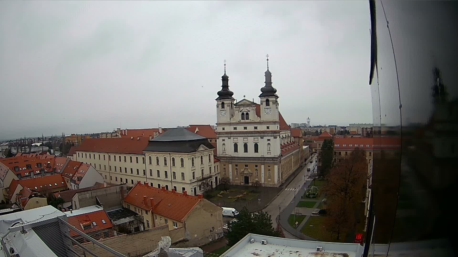 An overcast sky hangs over a historic European town, featuring a prominent baroque church with twin spires amidst a landscape of red-roofed buildings and city streets.