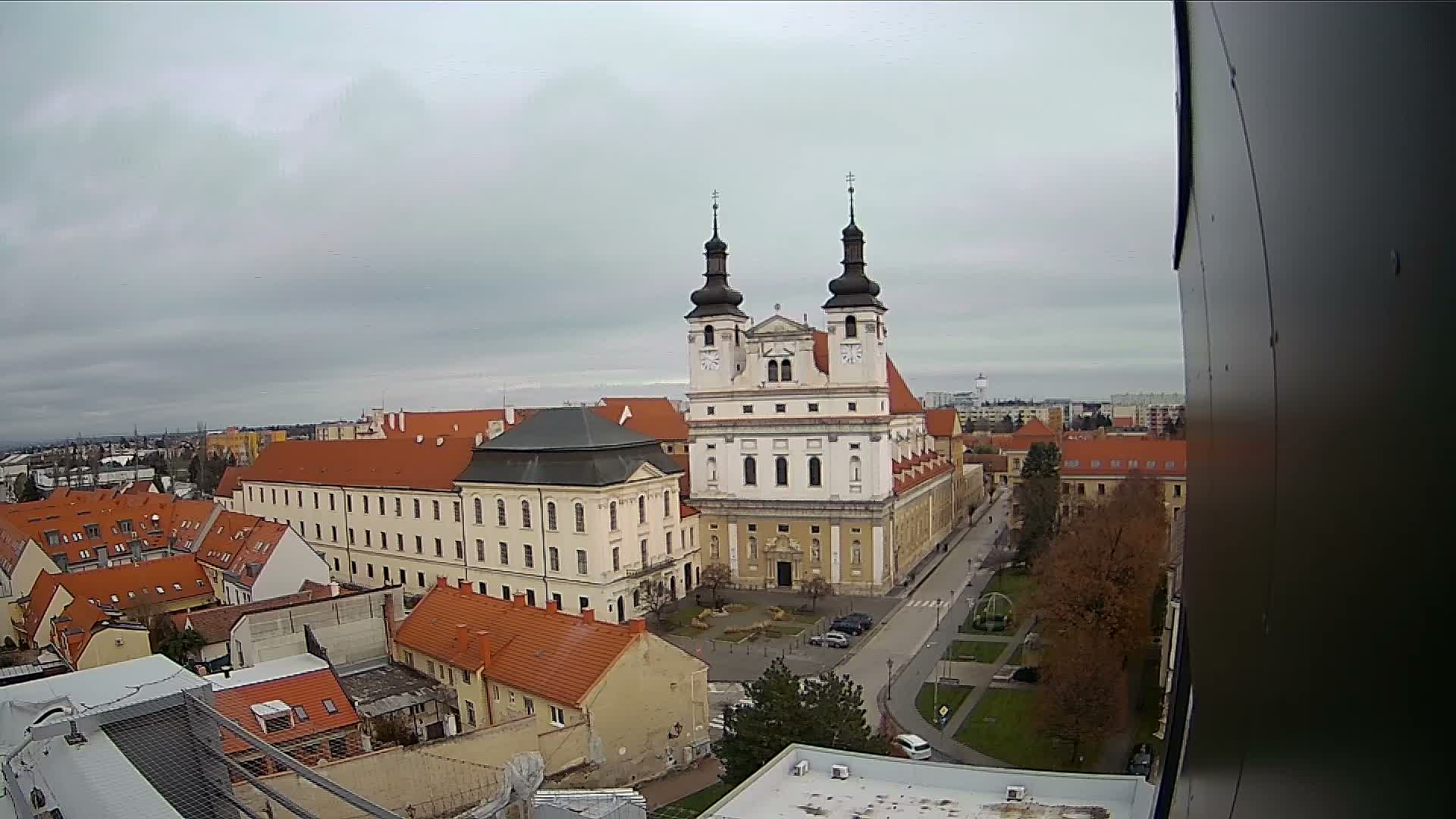 An elevated view reveals a historical European town dominated by a white church with twin dark steeples, surrounded by buildings with terracotta roofs and autumn trees, all under a grey, overcast sky.