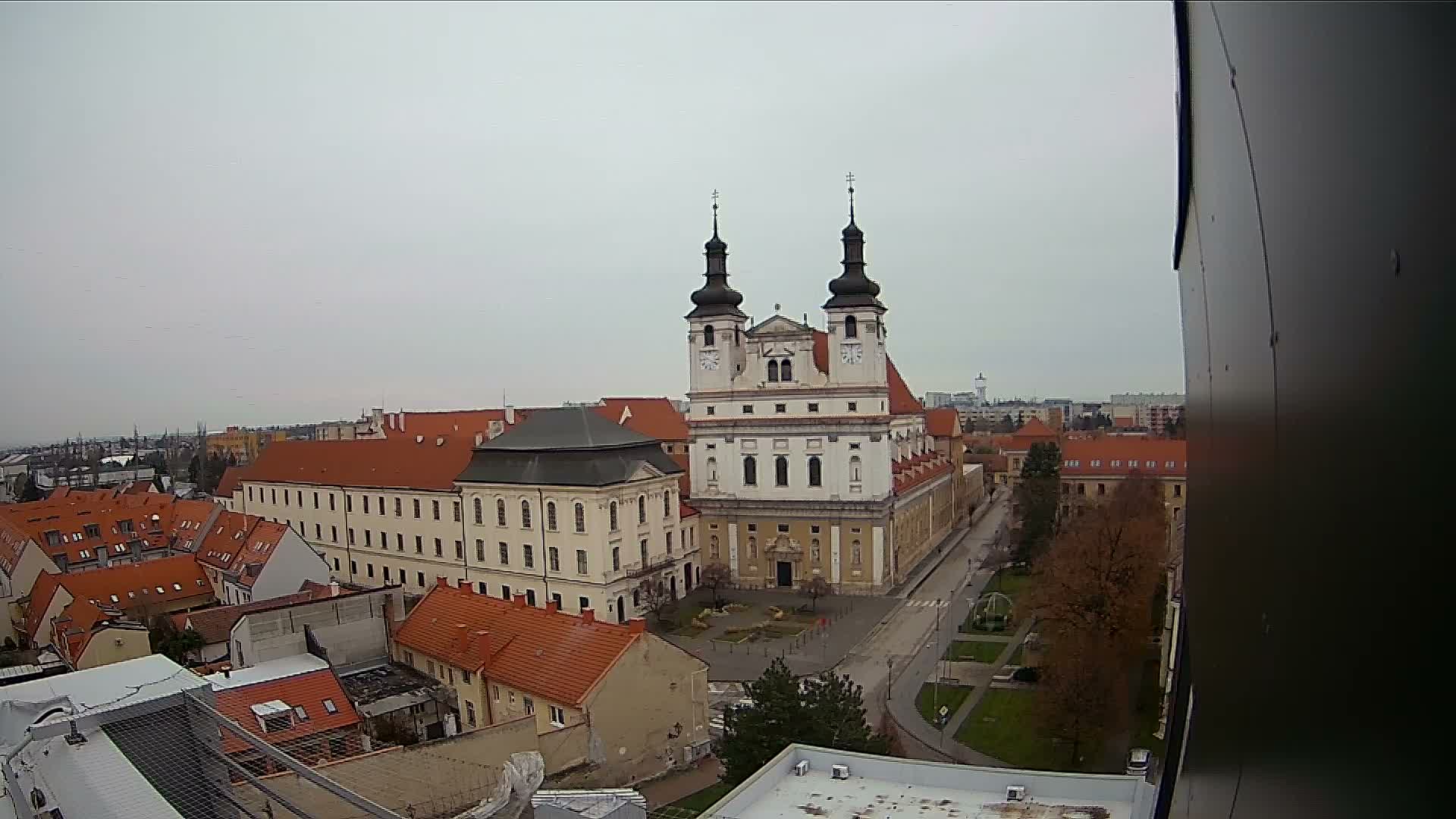 A prominent baroque church with two dark spires stands central in an overcast aerial view of a European town, surrounded by buildings with red roofs and autumn trees.
