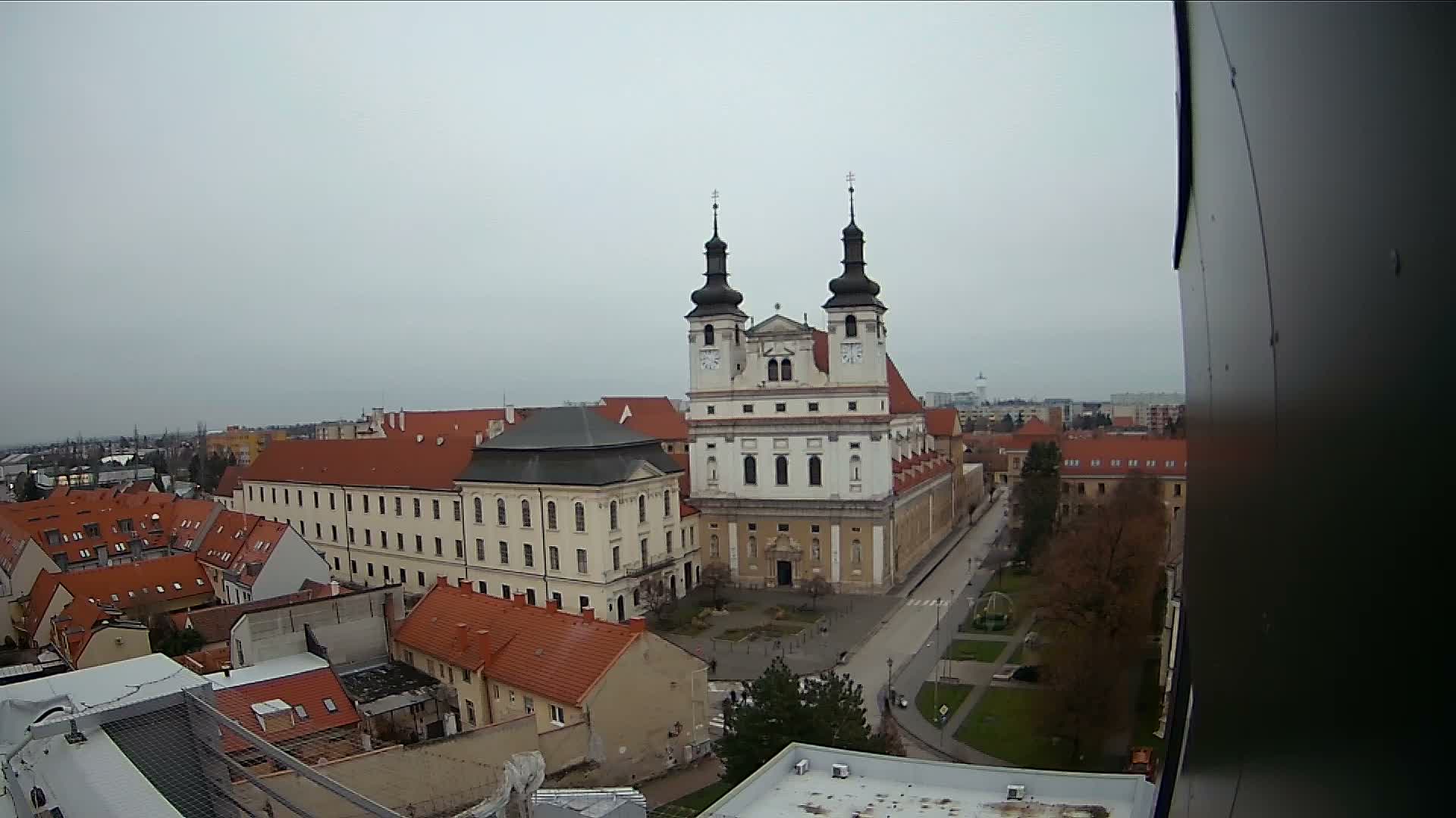 A prominent baroque church with two dark spires stands central in an overcast aerial view of a European town, surrounded by buildings with red roofs and autumn trees.