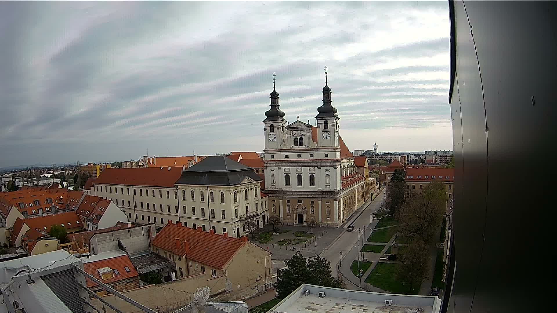 A prominent baroque church with two dark spires stands central in an overcast aerial view of a European town, surrounded by buildings with red roofs and autumn trees.