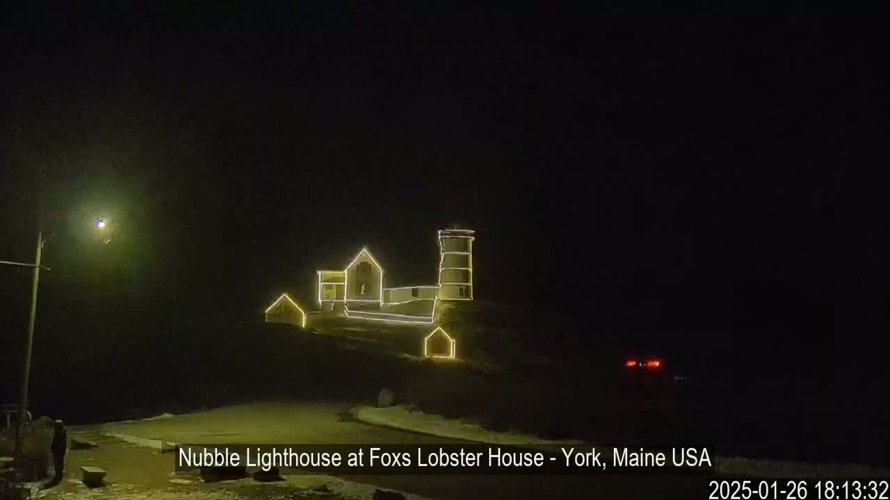 At night, a lighthouse and adjacent buildings are illuminated with yellow string lights.