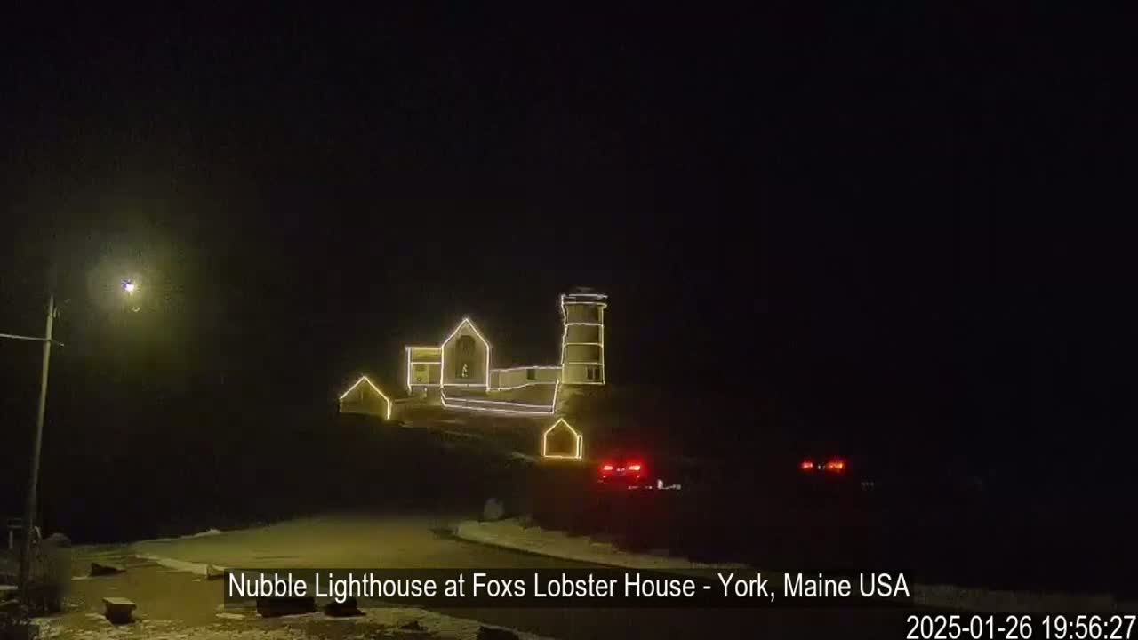 At night, a lighthouse and adjacent buildings are outlined with yellow string lights, with a few cars parked nearby on a snow-dusted ground.