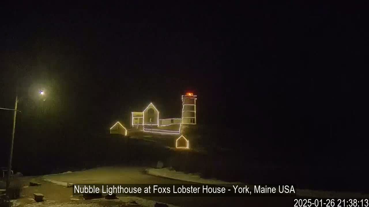 A lighthouse and adjacent buildings, outlined in yellow lights, are visible at night in what appears to be snowy conditions.