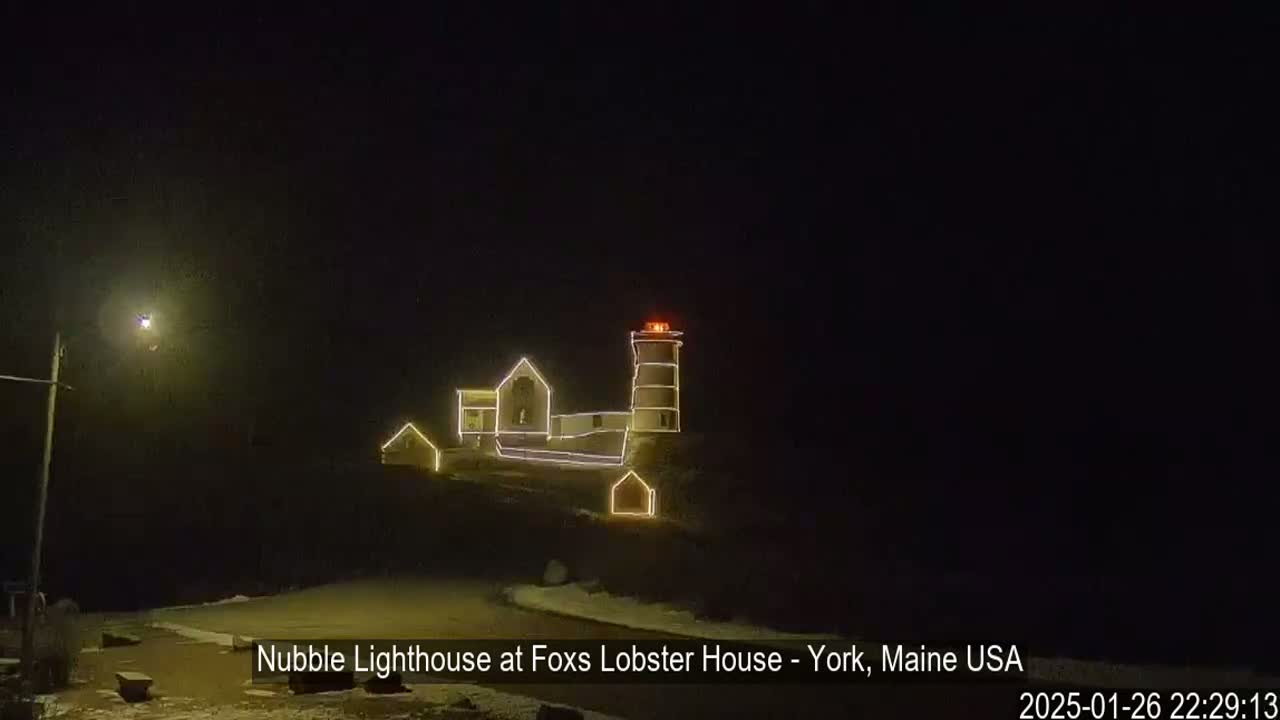 At night, a lighthouse and adjacent buildings are decorated with strings of lights, and a bit of snow is visible on the ground.