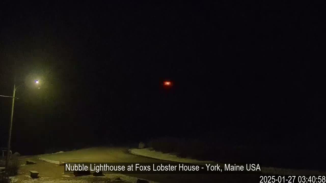A dark, nighttime scene shows a lit streetlight and a faint red light in the distance, with a snow-covered area visible in the foreground.