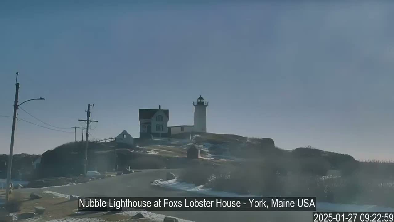A lighthouse and a smaller building sit atop a hill partially covered in snow under a clear, bright sky.