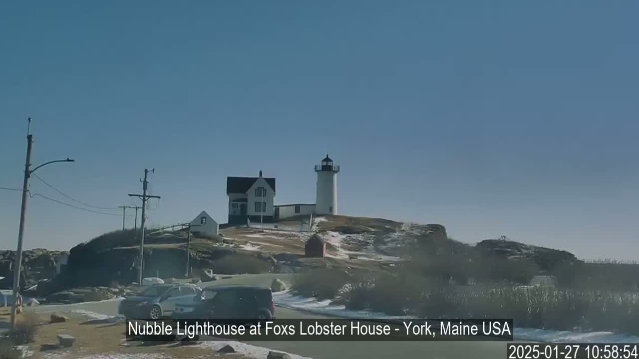 A white lighthouse and adjacent buildings sit atop a rocky hill partially covered in snow under a clear blue sky.
