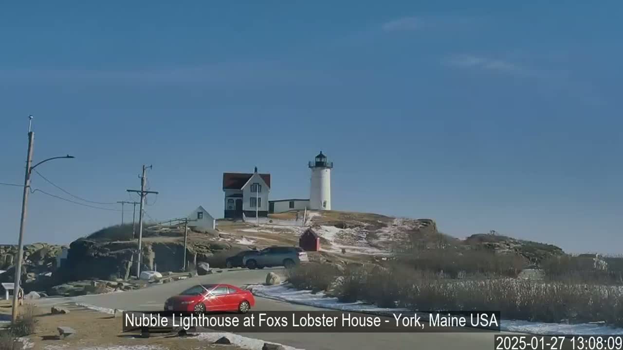 A white lighthouse and adjacent building are situated on a rocky outcrop with patches of snow under a clear blue sky.