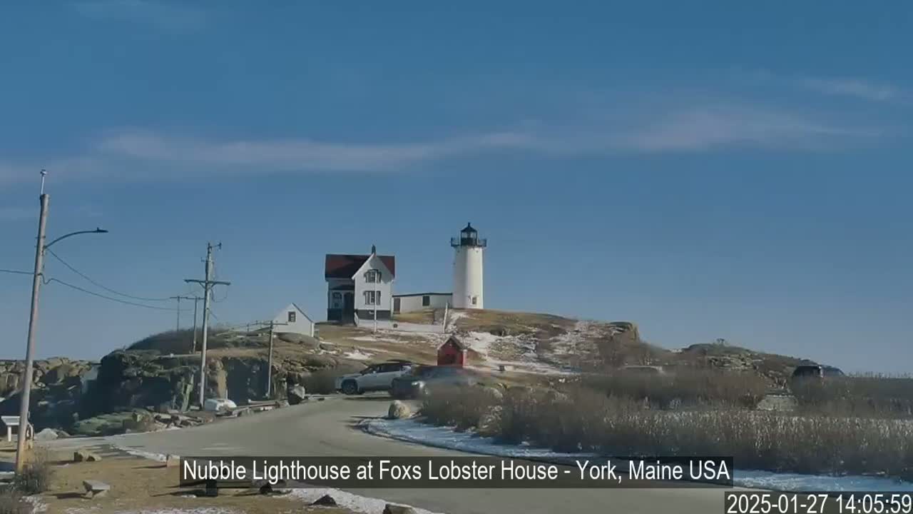 A white lighthouse and a house sit atop a rocky hill overlooking a road with a few cars parked, under a clear blue sky with a few wispy clouds and patches of snow on the ground.