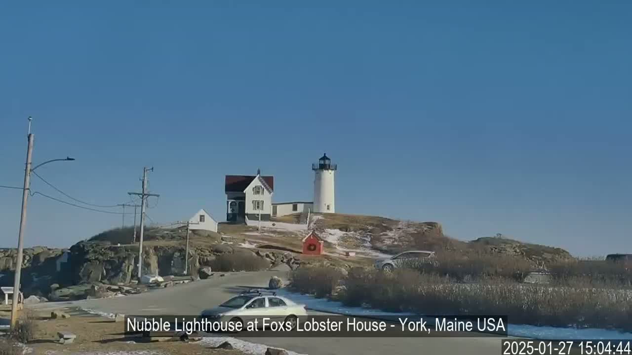 A lighthouse and a house stand on a rocky hill overlooking a parking lot with cars, under a clear blue sky on a partly snowy day.