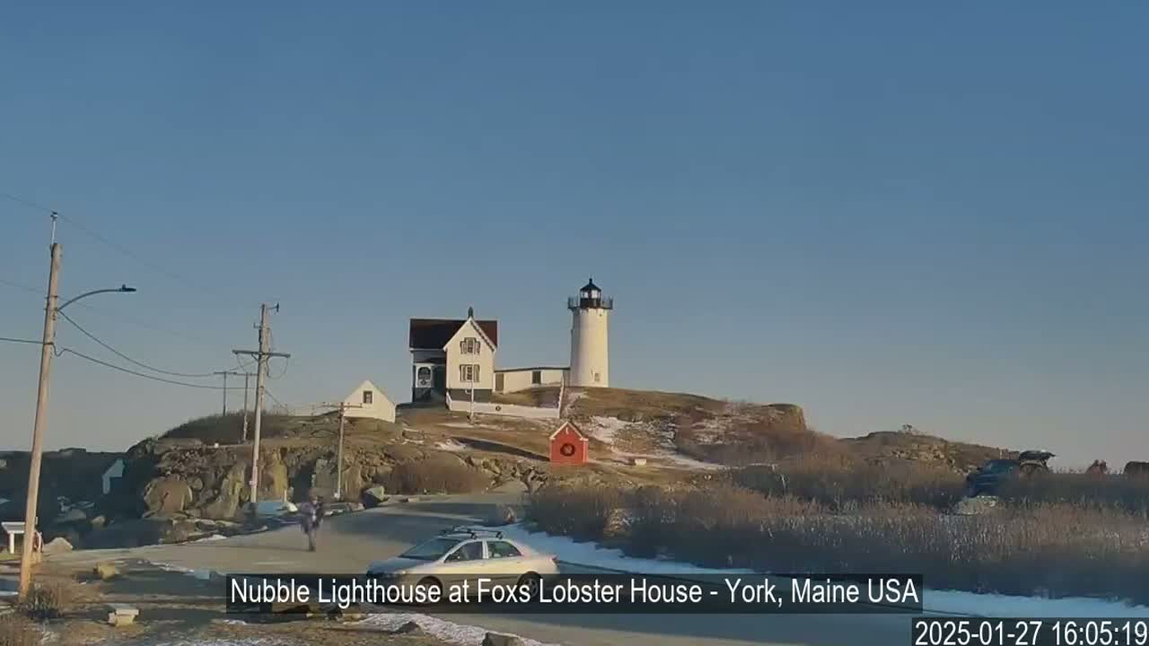 A white lighthouse sits atop a rocky outcrop, next to a smaller building, under a clear blue sky on a partly snowy day.