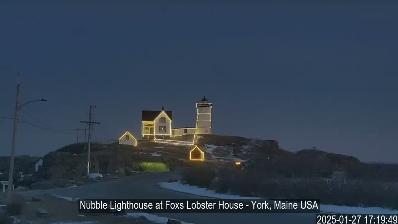 A lighthouse and keeper's house, adorned with string lights, sit atop a snow-dusted hill under a twilight sky.