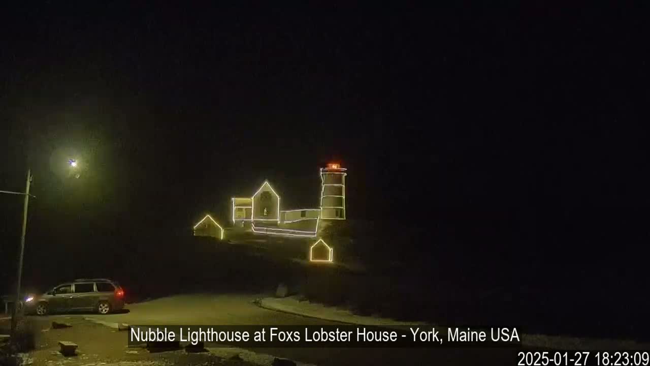 A nighttime view of a lighthouse and adjacent buildings decorated with string lights, a vehicle parked nearby under a dark sky.