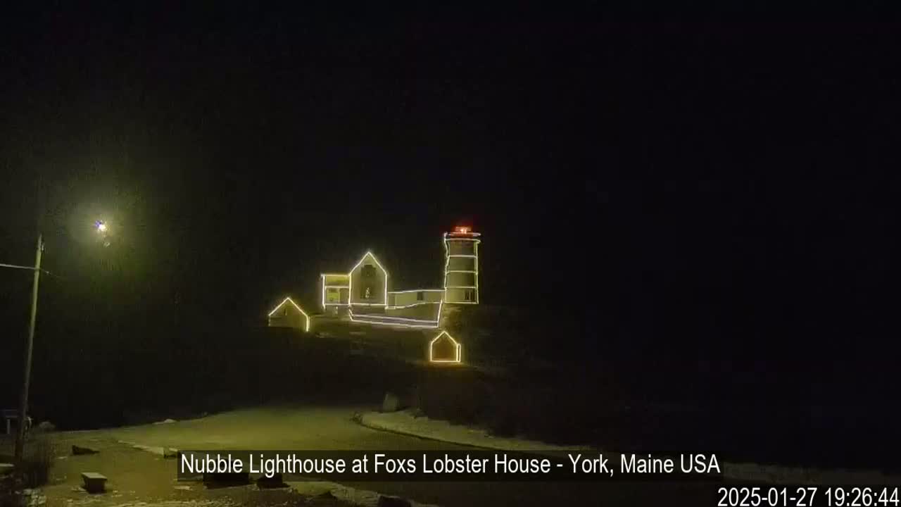 A lighthouse and adjacent buildings are illuminated with string lights at night in snowy conditions.