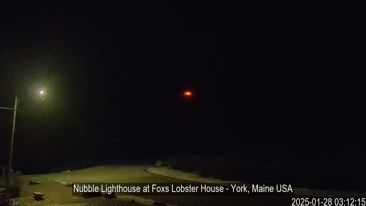 A nighttime scene shows a lighthouse in the distance, illuminated by its own light, with a streetlight visible in the foreground under a dark, possibly snowy sky.