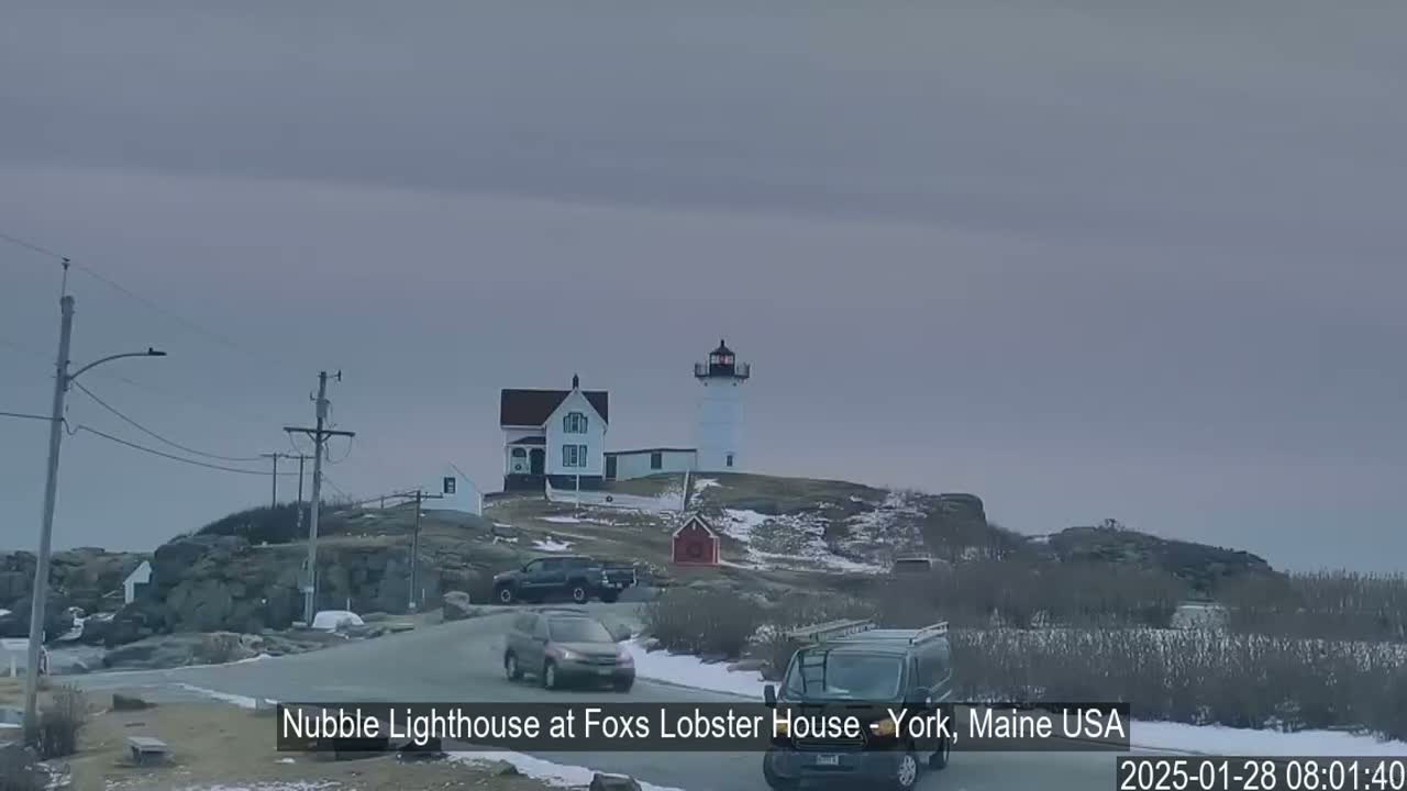 A lighthouse and keeper's house sit atop a rocky, snow-dusted hill, with several vehicles on a road in the foreground under an overcast sky.