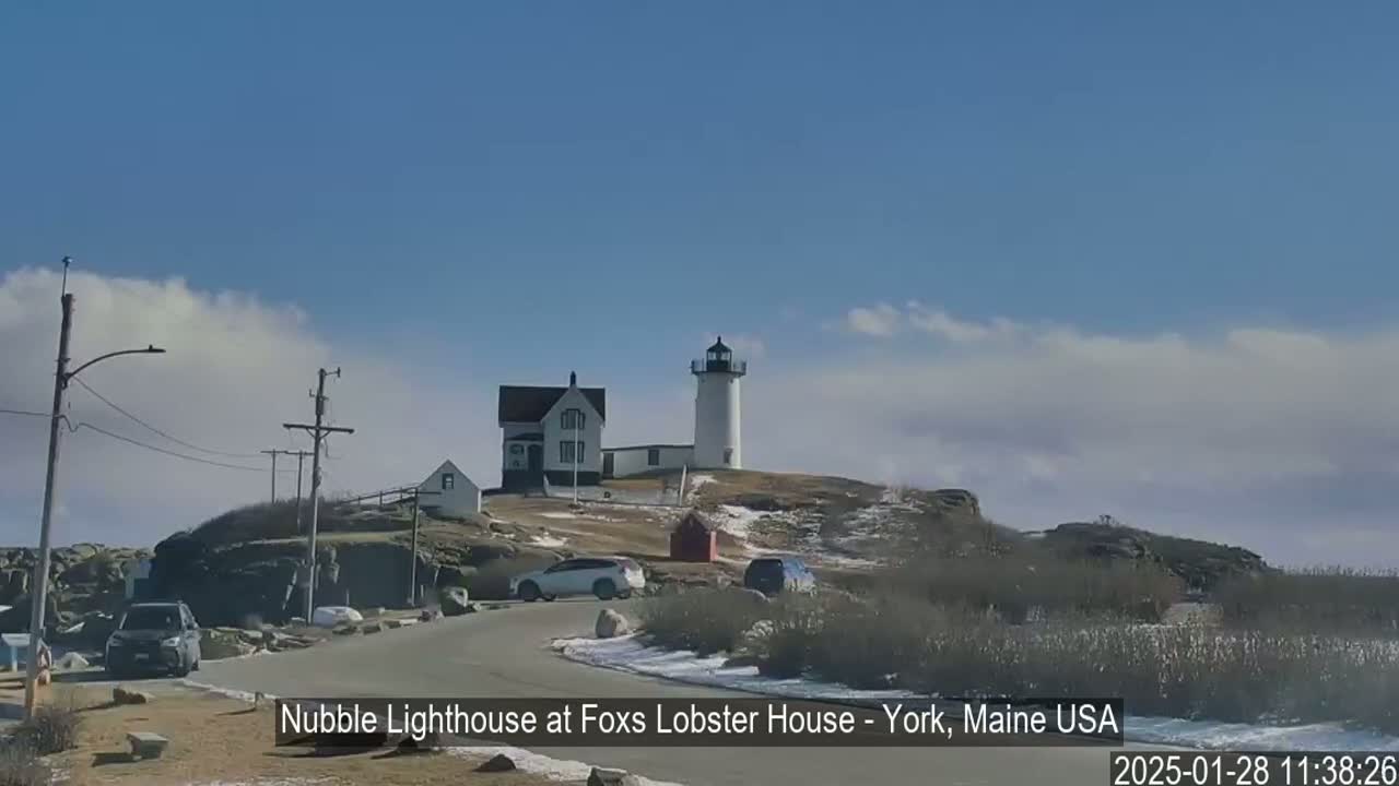 A white lighthouse sits atop a rocky hill with a few cars parked nearby, under a mostly sunny sky with some clouds.
