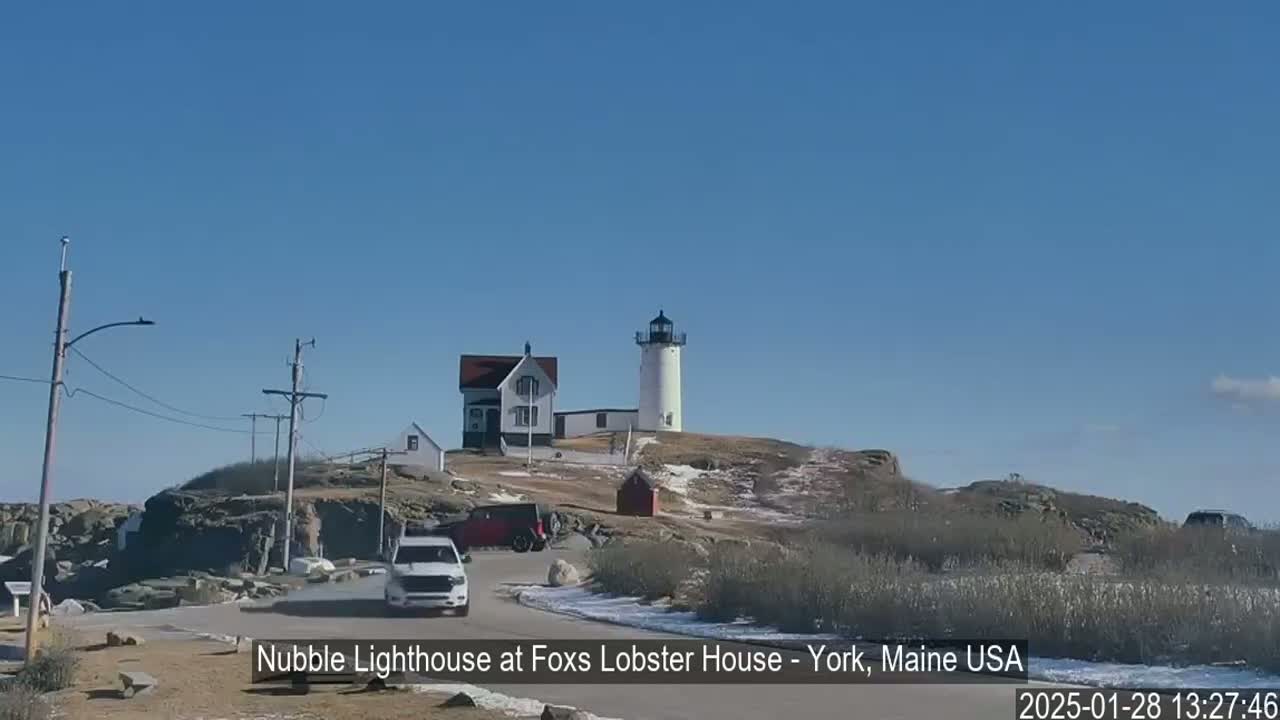 A lighthouse and keeper's house sit atop a rocky hill overlooking a road with a few vehicles, under a clear blue sky with patches of snow on the ground.