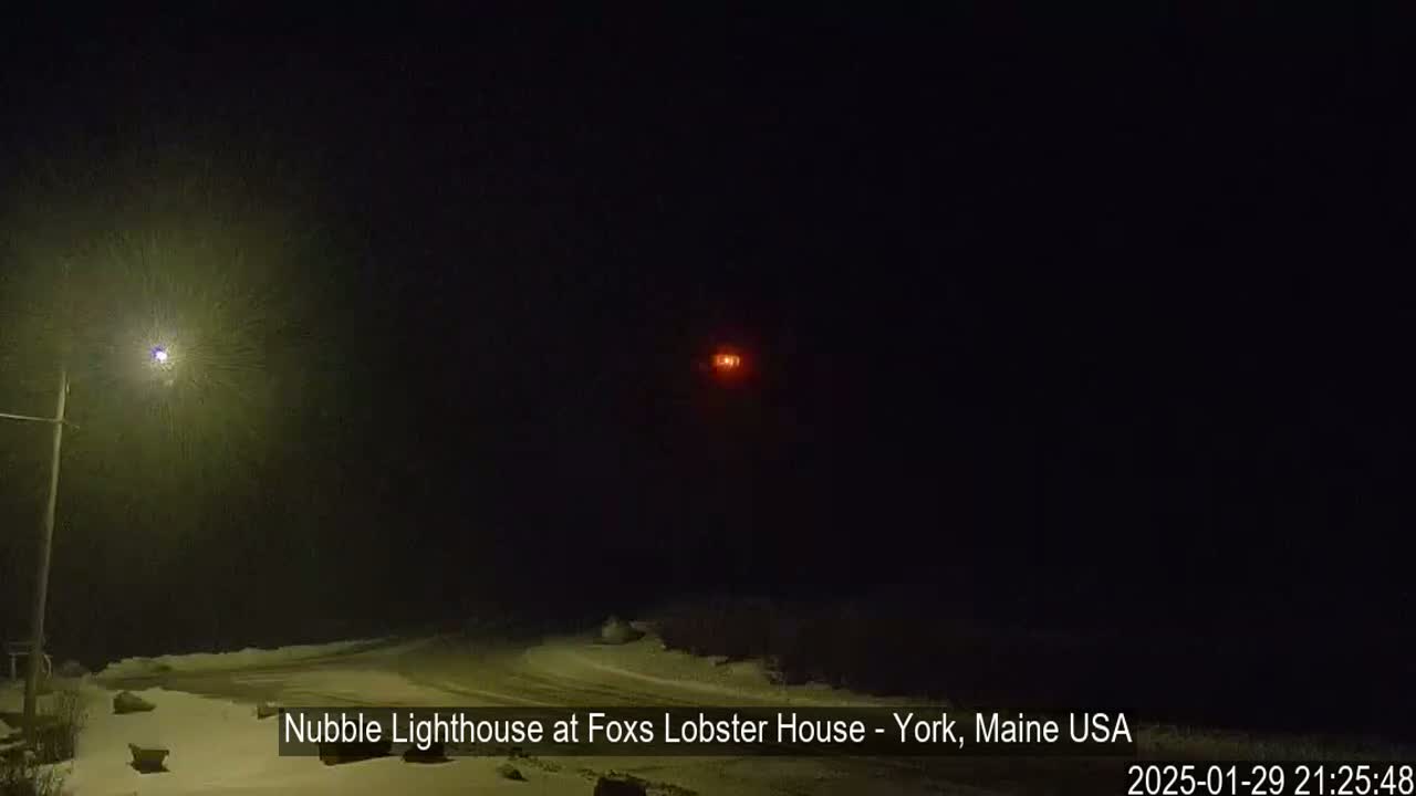 A snowy, dark nighttime scene shows a lighthouse's light in the distance, illuminated by a nearby streetlight.