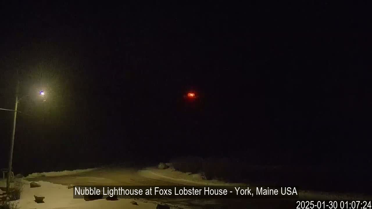 A snow-covered landscape is illuminated at night by a streetlight and a distant red light, possibly a lighthouse.