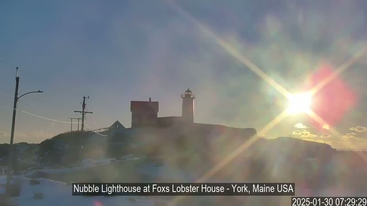 A lighthouse and adjacent buildings are silhouetted against a bright, hazy sunrise, with a dusting of snow on the ground.