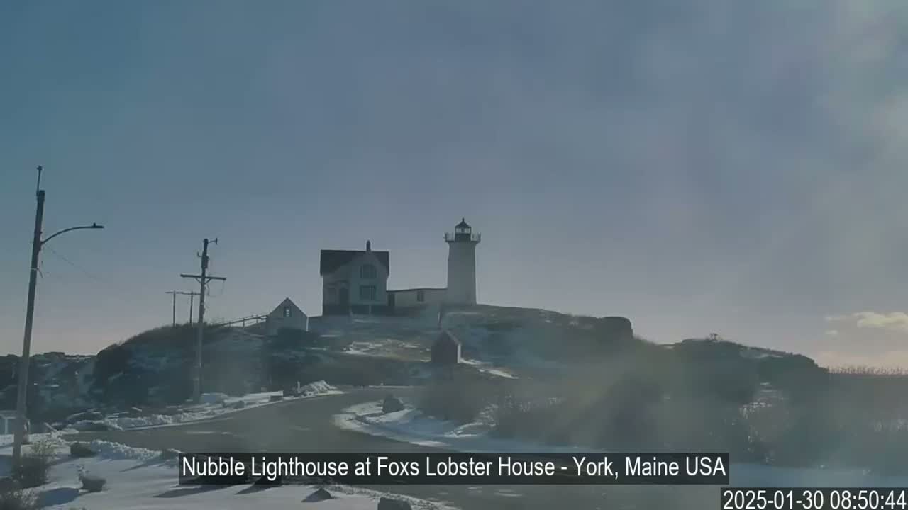 A lighthouse and adjacent buildings sit atop a snow-dusted, rocky hill under a clear, bright sky.
