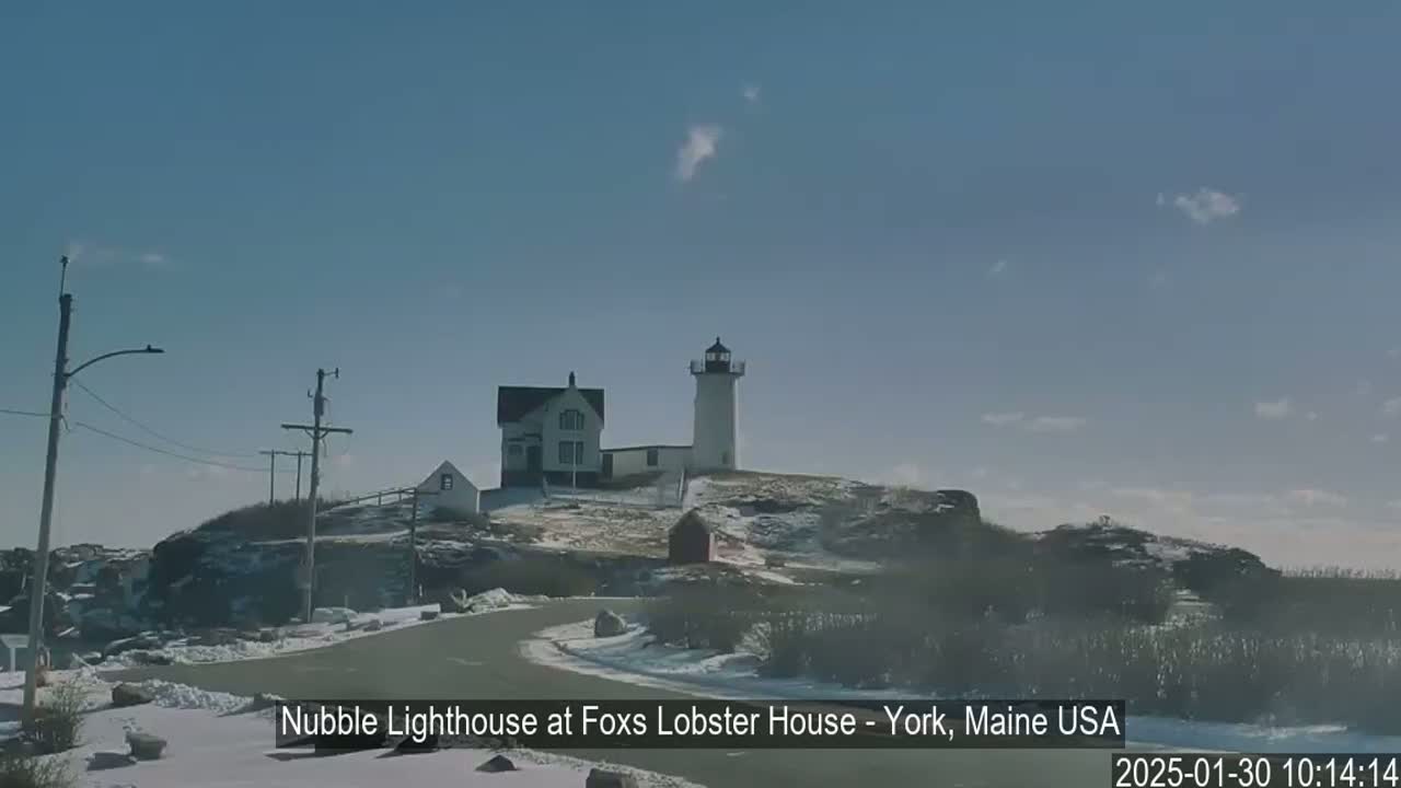 A lighthouse and other buildings sit atop a snow-covered rocky hill on a partly cloudy day.
