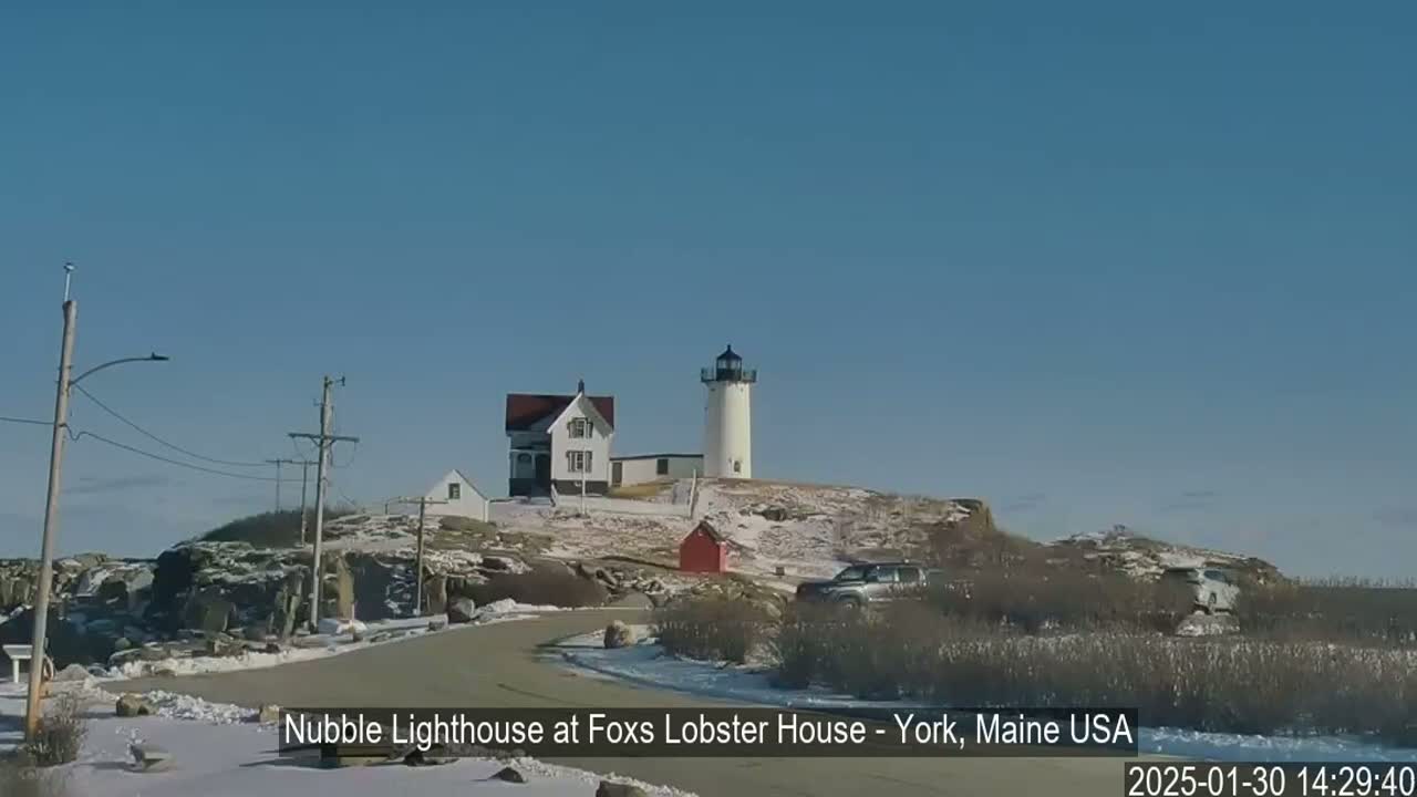 A white lighthouse and a smaller white building sit atop a snow-dusted rocky hill on a sunny day.