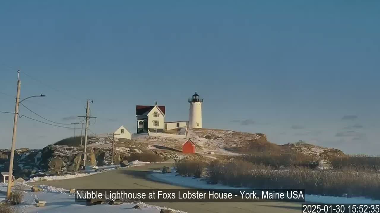 A lighthouse and other buildings sit atop a snow-dusted rocky hill under a clear blue sky.