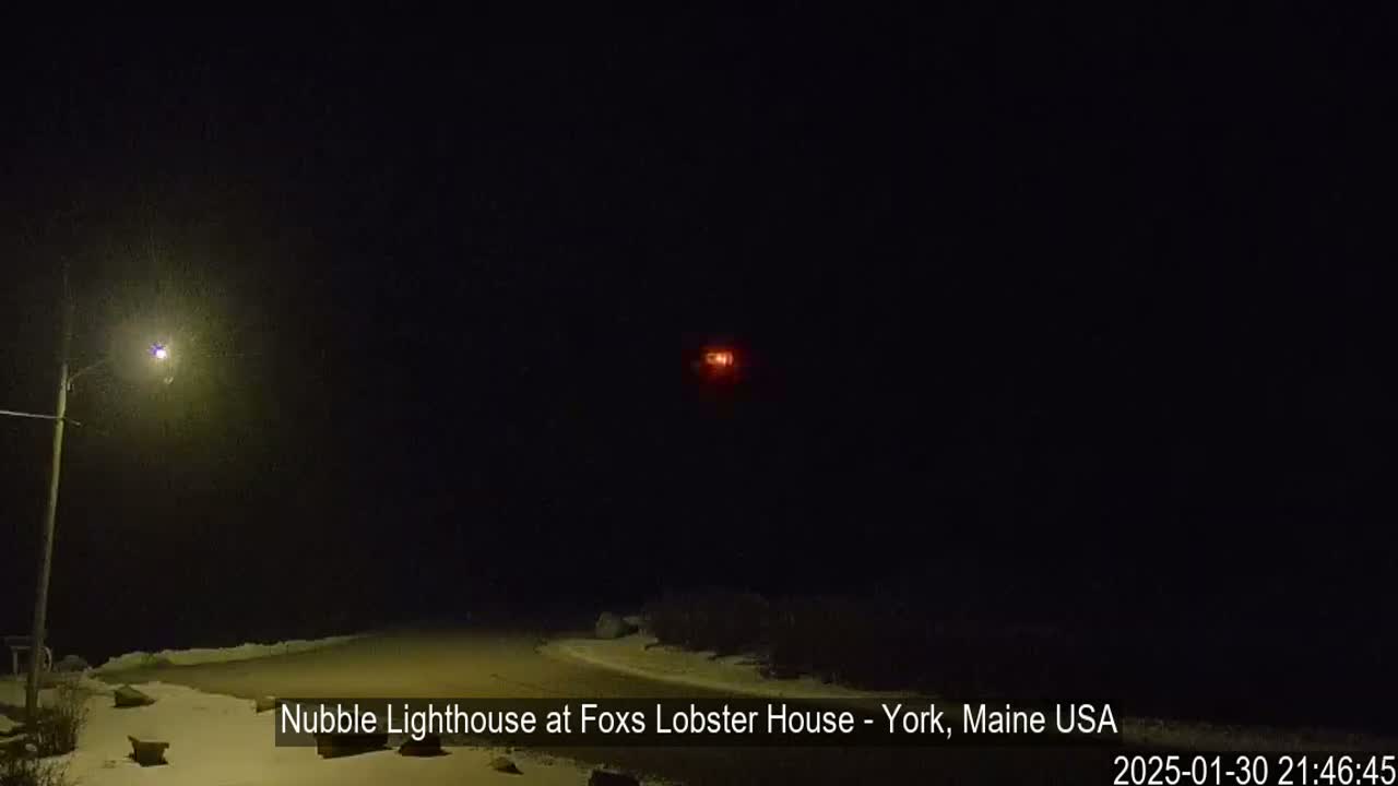 A nighttime view of a snow-covered area with a distant, faint red light and a lit streetlamp.