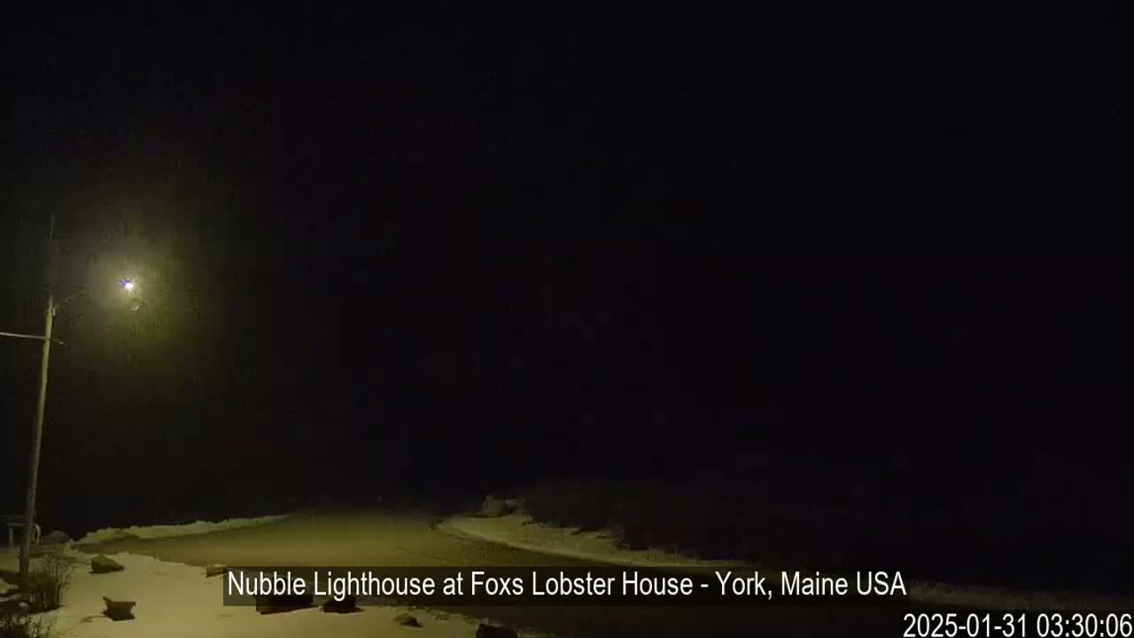 A nighttime, snowy scene shows a dimly lit roadway near a coast, with a single lamppost visible.