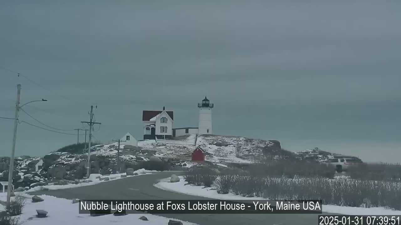A lighthouse and a house sit atop a snow-covered rocky hill under an overcast sky.