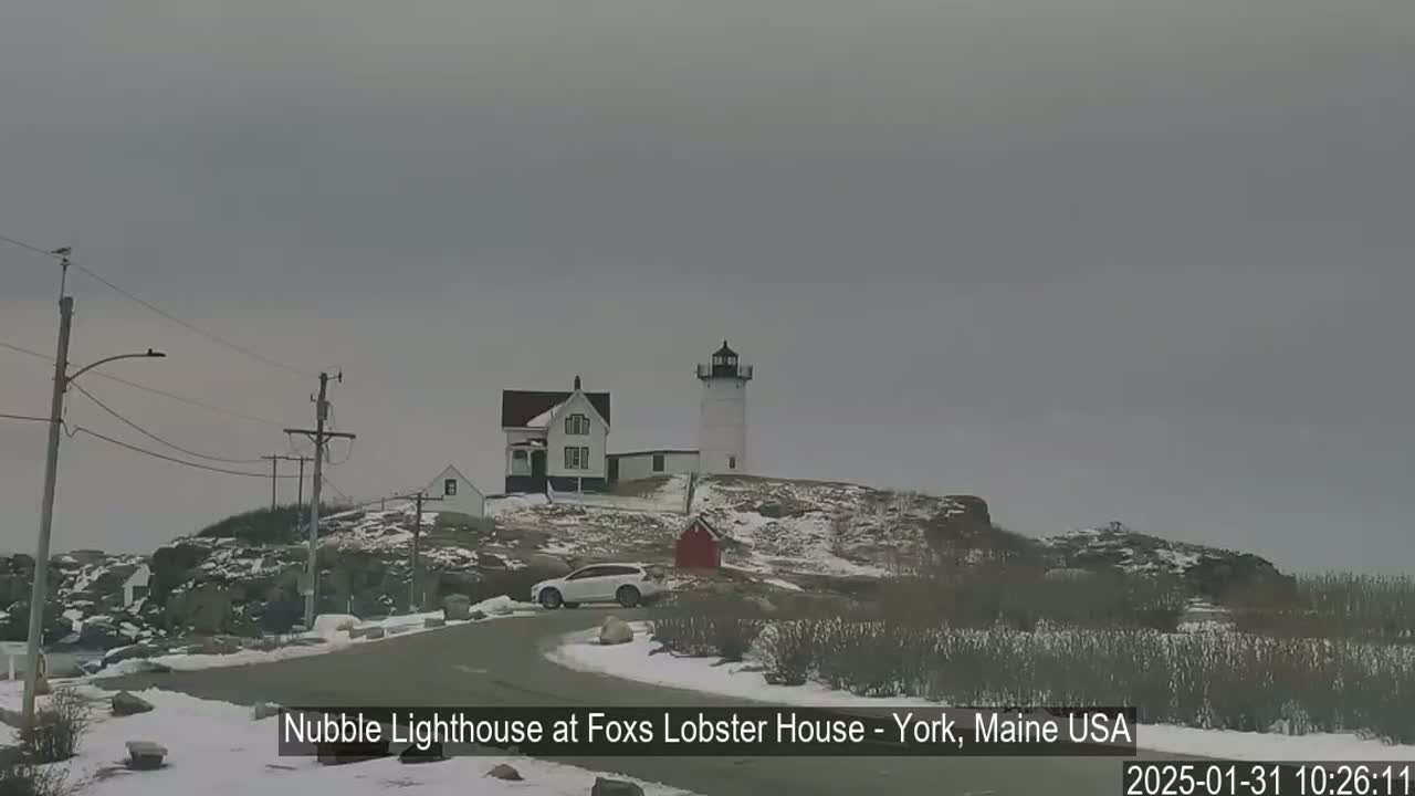 A white lighthouse and a house sit atop a snow-covered, rocky hill under an overcast sky.