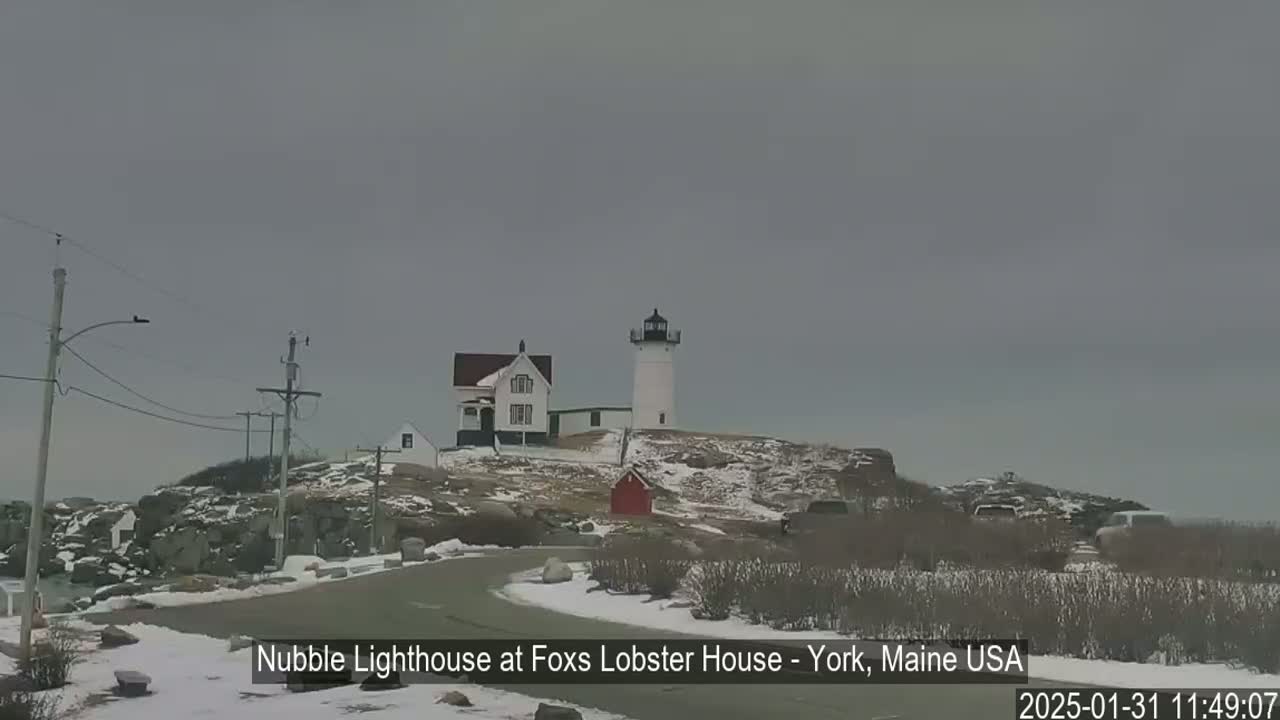 A white lighthouse and keeper's house are situated on a snow-covered rocky outcrop under an overcast sky.